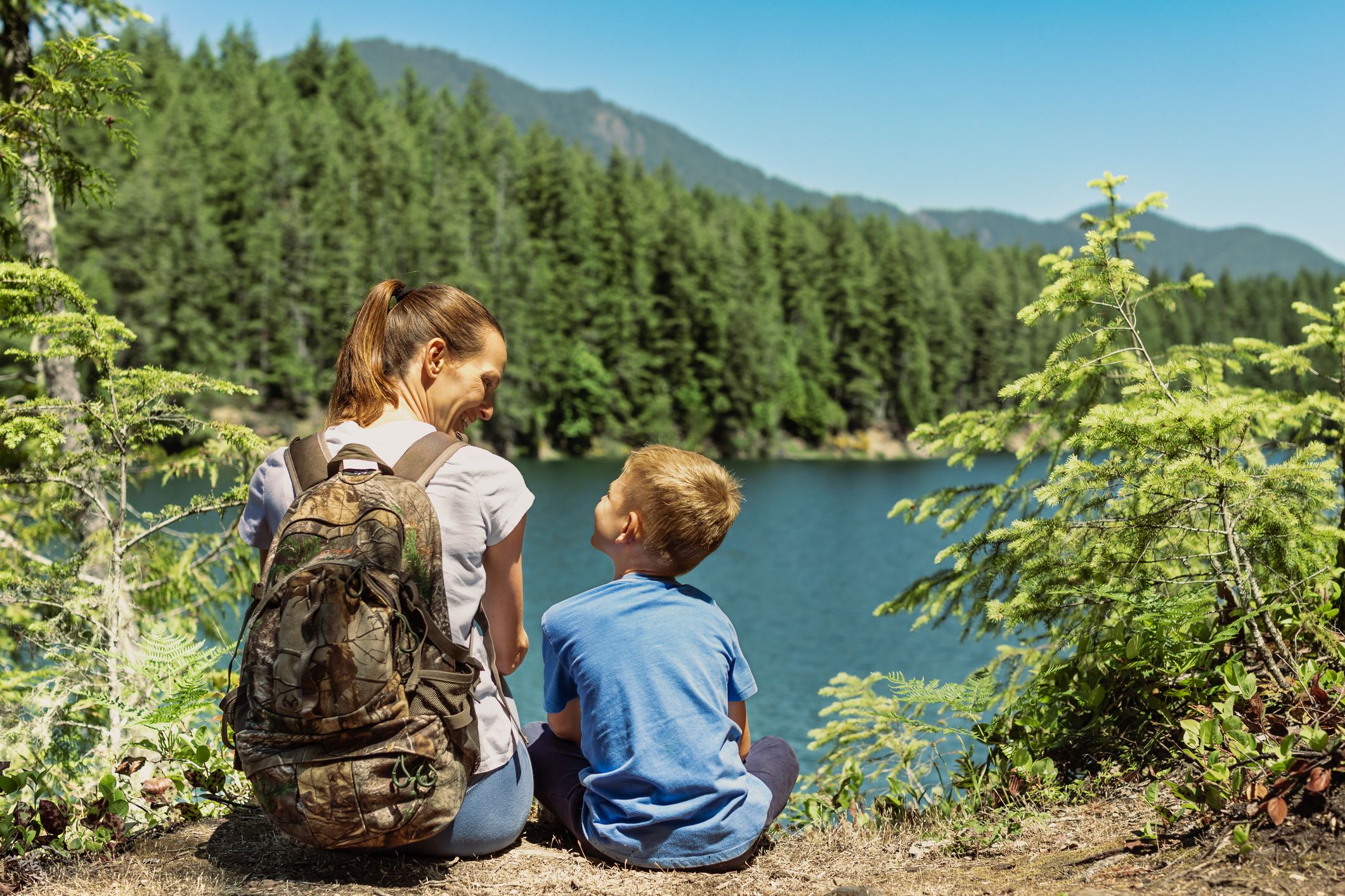 A woman wearing a backpack and a young boy smiling at each other while sitting in the woods next to a lake