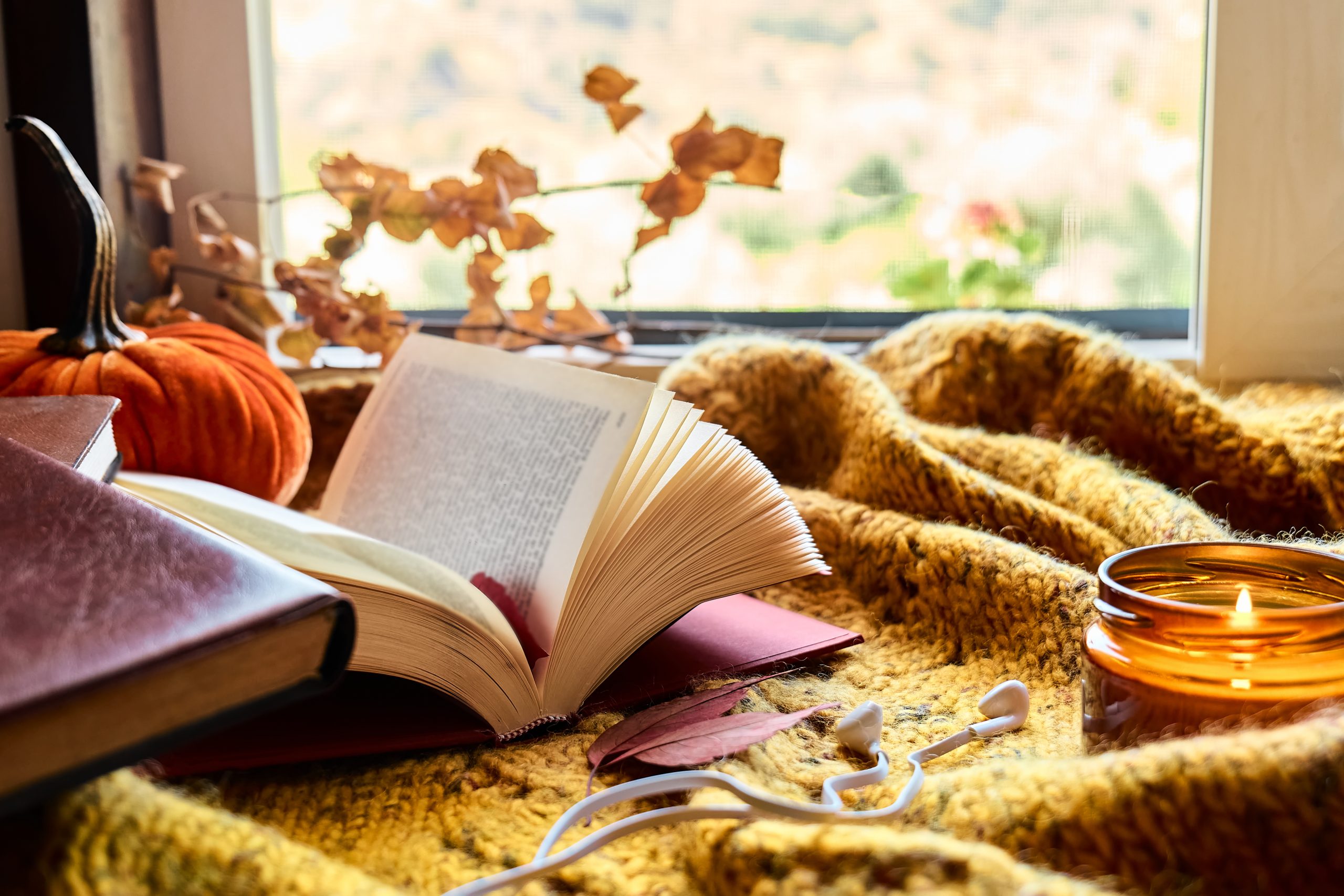 Cozy indoor fall scene with rust-colored leaves, blanket, books, a lit candle, a plush stuffed pumpkin decoration, and earbuds.