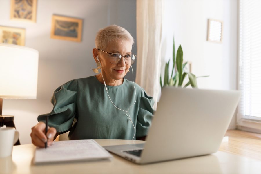 Woman wearing headphones writing on her notepad while watching something on her laptop